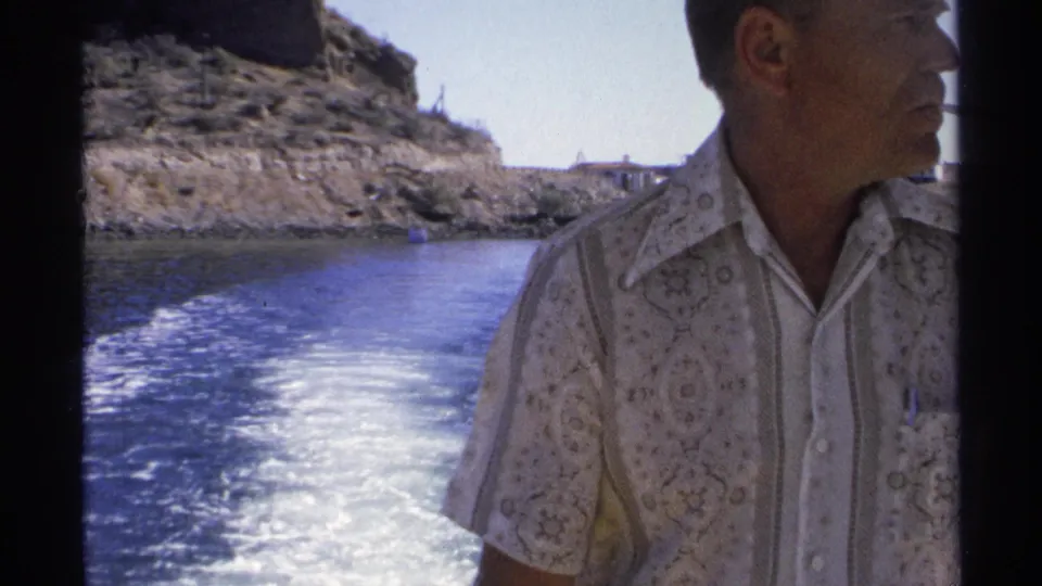 A man is partly framed at the side of a moving boat, with the Guaymas shoreline and foamy wake visible behind him.