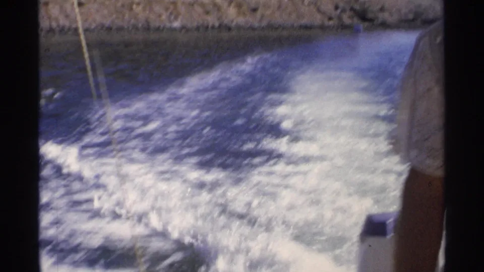 The moving boat's wake fills most of the frame, with a partial passenger and rocky Guaymas shoreline at the margins.