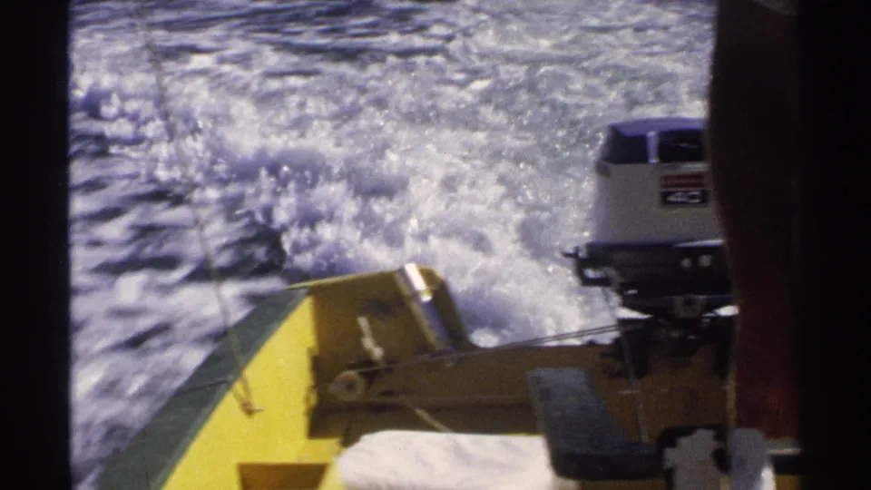 Rear-facing 1975 Guaymas frame with a white-and-blue outboard motor, yellow stern, and foamy wake behind the boat.