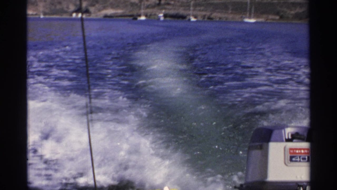 1975 Guaymas home-movie frame showing blue water, white wake, a Mercury outboard motor, and anchored sailboats near rocky hills.