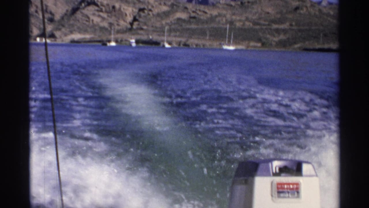 Boat wake spreads across blue water behind a visible outboard motor near anchored sailboats.