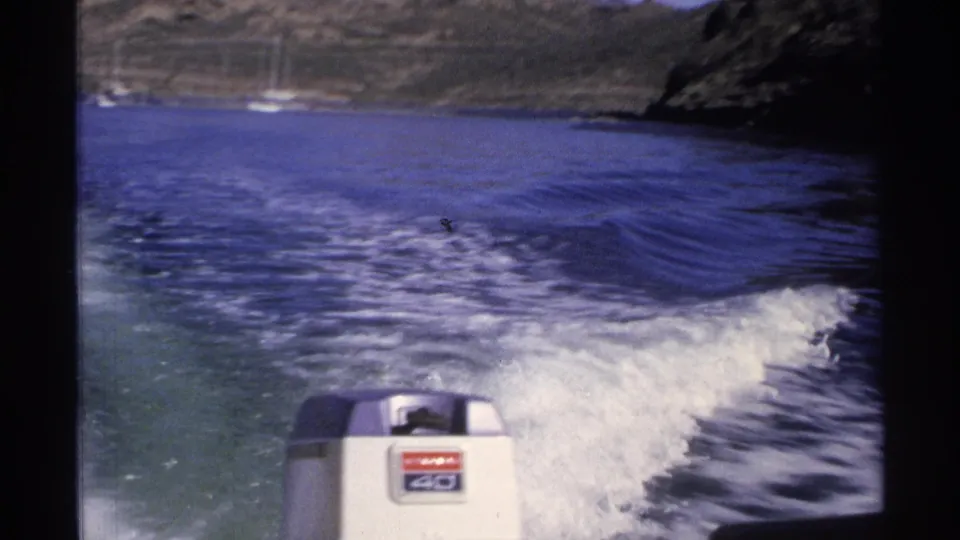 1975 8mm frame from Guaymas showing a Mercury outboard motor, churning wake, blue water, and distant sailboats near the coast.