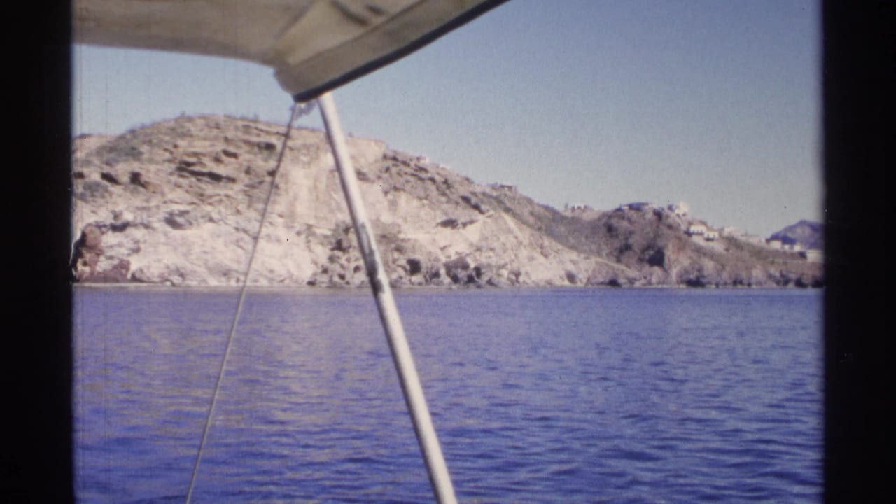 1975 Guaymas, Mexico view from a boat, with calm sea, pale rocky hills, distant buildings, and a canopy support in frame.