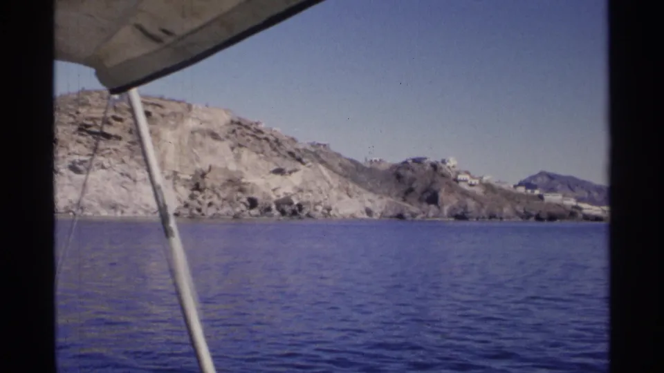 Boat-framed view of Guaymas coastline in 1975, with rippled blue water, rocky slopes, and small white buildings in the distance.