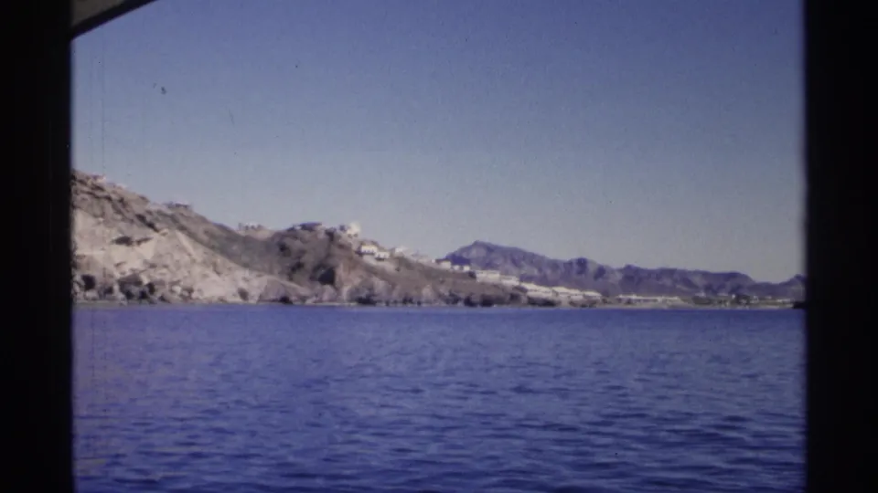 Wide 1975 Guaymas shoreline view from the water, with rocky hills, blue sea, distant white buildings, and mountains beyond.