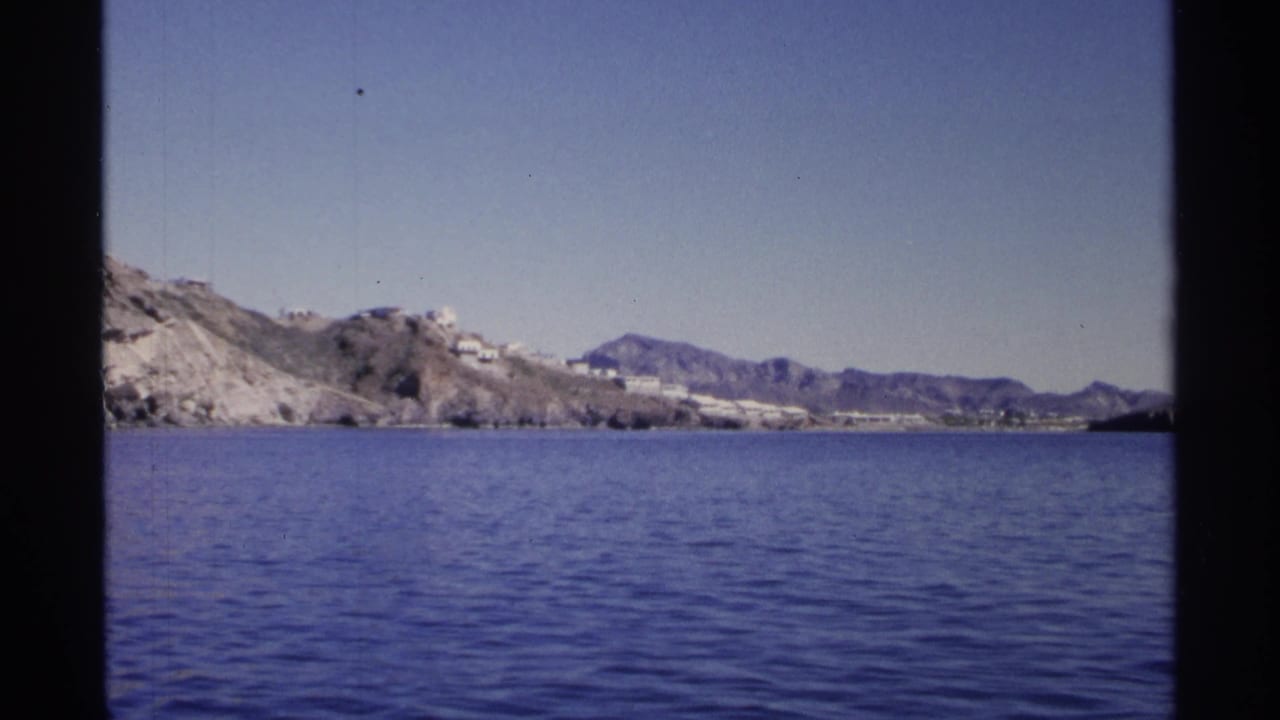 1975 8mm view likely near Guaymas, Mexico, showing blue water, arid shoreline, white hillside buildings, and distant mountains.