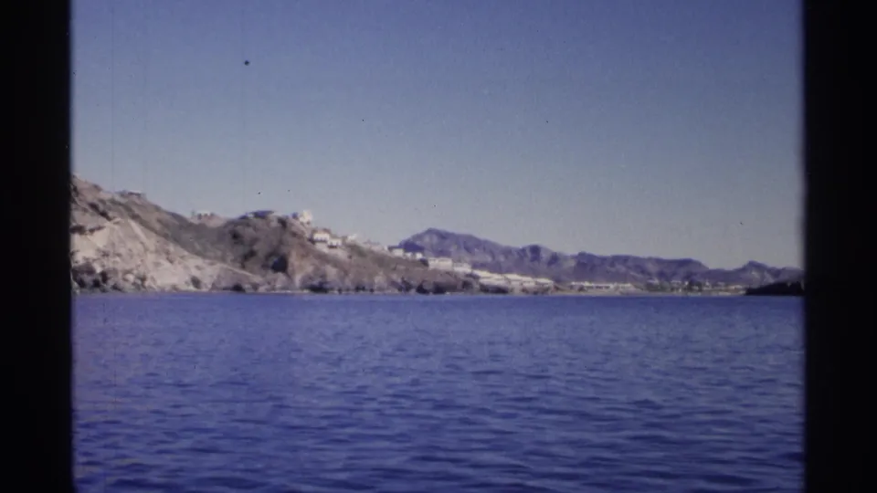 1975 8mm view likely near Guaymas, Mexico, showing blue water, arid shoreline, white hillside buildings, and distant mountains.