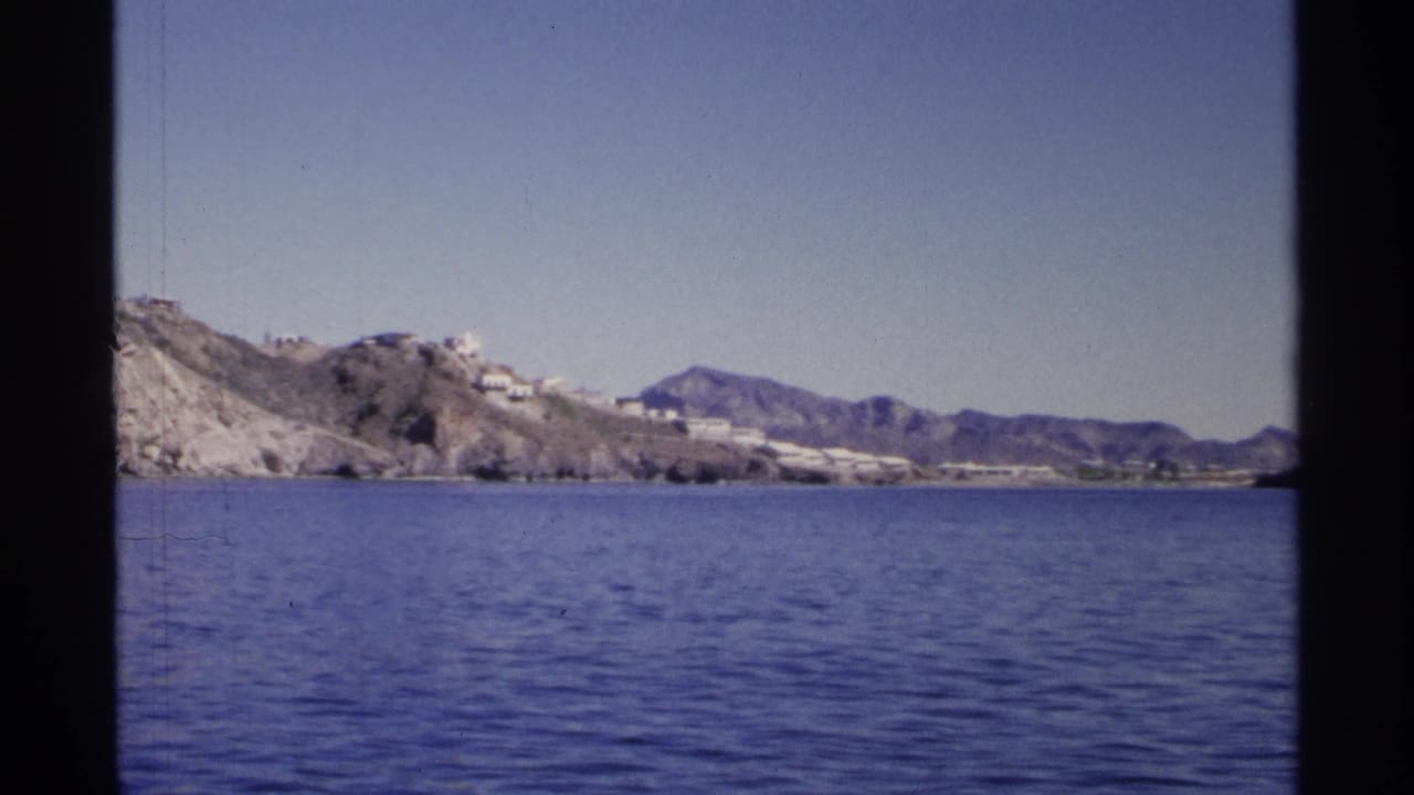 Wide coastal view with water, rocky hills, white buildings, and mountains under clear sky.