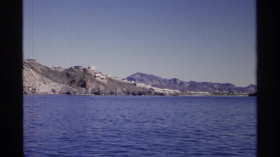 Likely Guaymas, Mexico in 1975, seen from the water with arid hills, pale buildings, and mountains across the shoreline.
