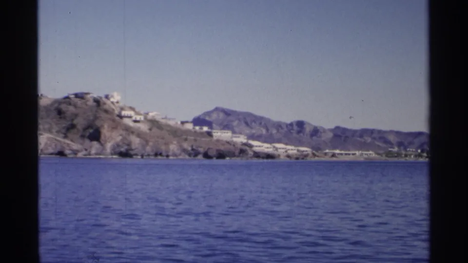 1975 home-movie frame likely near Guaymas, showing blue water below a rocky coast with white buildings and mountains behind.