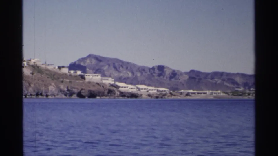 Likely Guaymas, Mexico, 1975, with calm water in front of rocky coastal land, white structures, and a broad mountain ridge.