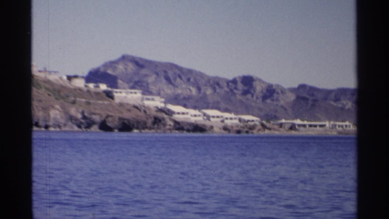 1975 Guaymas, Mexico home-movie frame showing white waterfront buildings below rugged mountains across calm blue water.