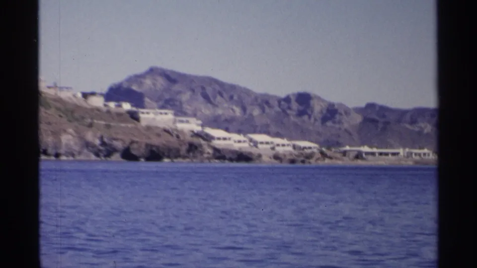 1975 Guaymas, Mexico home-movie frame showing white waterfront buildings below rugged mountains across calm blue water.