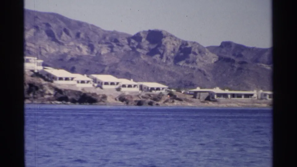 1975 Guaymas waterfront view with low white buildings, rocky terrain, mountains, and calm water in a soft 8mm frame.