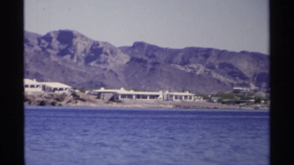 Final 1975 Guaymas frame with low buildings on a rocky waterfront, blue water foreground, and rugged mountain backdrop.