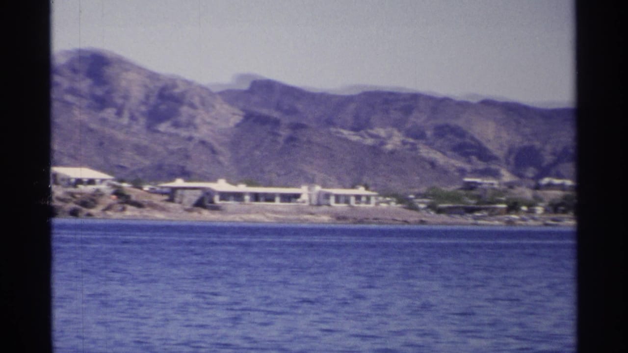 1975 Guaymas home-movie frame showing blue water, low shoreline buildings, pale sky, and arid mountains in the background.