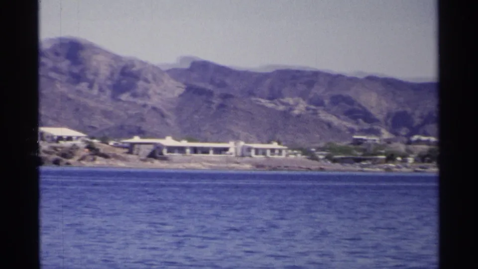 1975 Guaymas home-movie frame showing blue water, low shoreline buildings, pale sky, and arid mountains in the background.