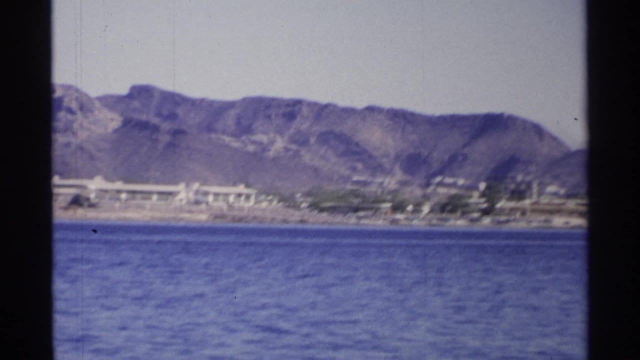 Mountain ridge and low shore buildings seen across blue water.