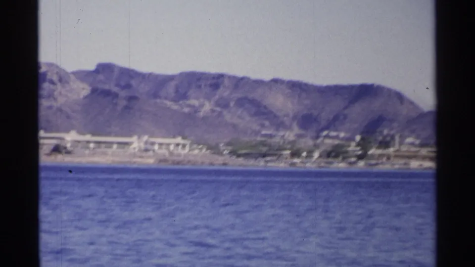 1975 Guaymas shoreline view with calm water in the foreground, low buildings near the coast, and brown mountain ridges behind.