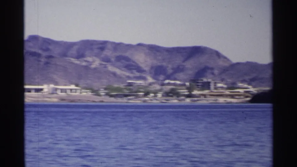 Soft 8mm frame of the Guaymas coast with blue water, low shoreline structures, and rugged mountains under a pale sky.