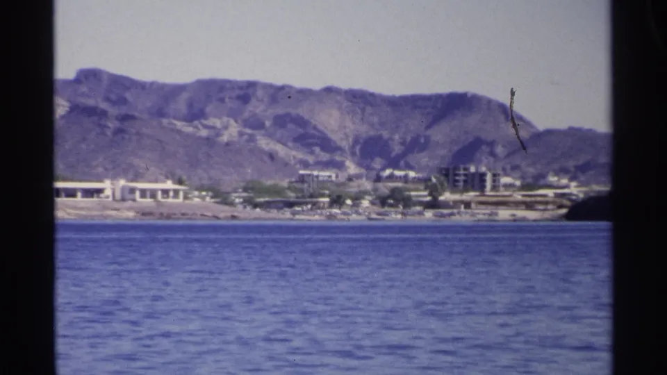 1975 Guaymas waterfront frame showing blue water, scattered shoreline buildings, a dark land edge, and rugged mountains behind.