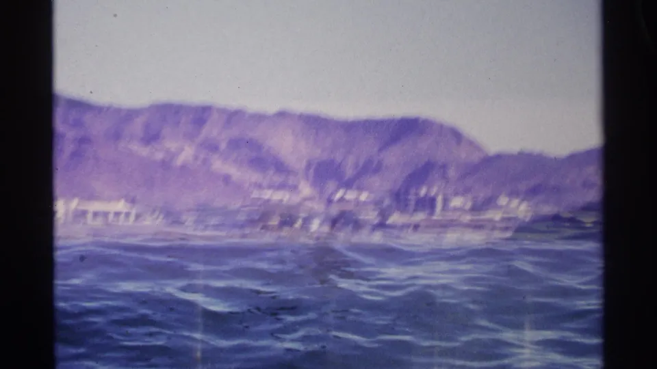 1975 Guaymas home-movie frame showing blue water, distant shoreline buildings, and mountains beyond the harbor.