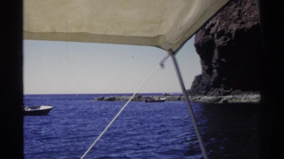 1975 Guaymas, Mexico 8mm frame looking from under an awning toward blue sea, a small boat, and a steep rocky coast.
