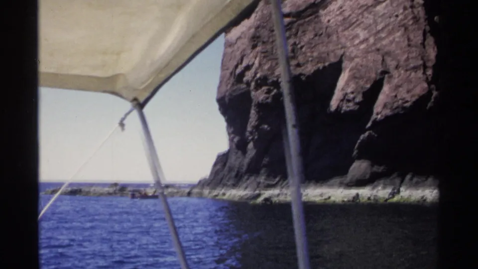 1975 Guaymas, Mexico frame showing rugged coastal rock, blue water, and the filming boat's awning and pole in the foreground.