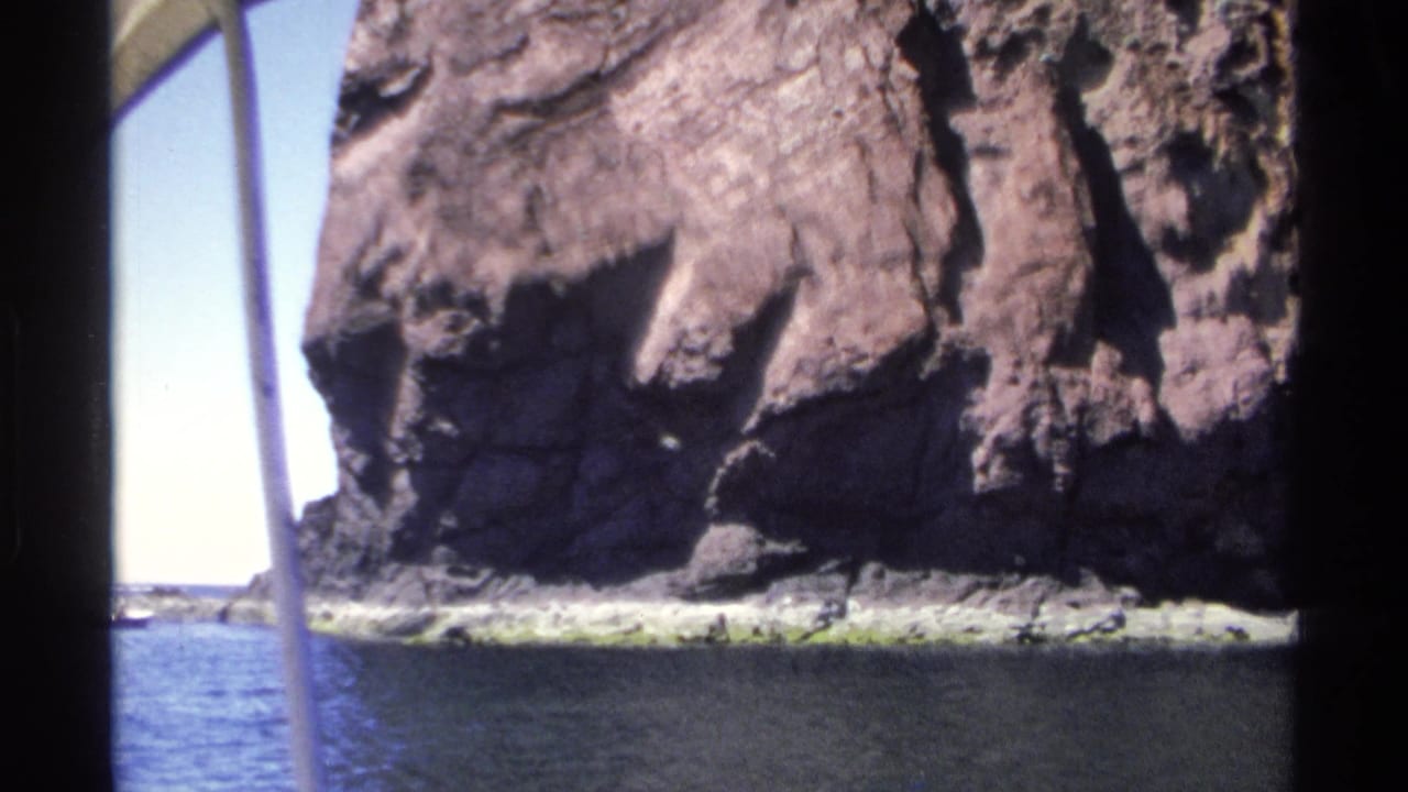 Rock cliff and narrow shoreline seen across blue water from a boat.