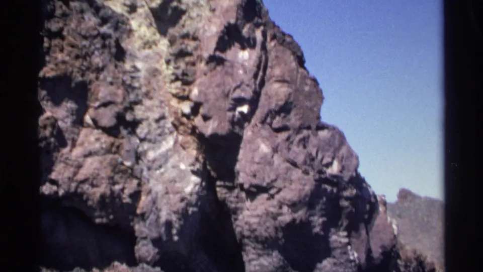 1975 Guaymas home-movie frame showing jagged coastal rock formations with shadowed crevices and pale blue sky.