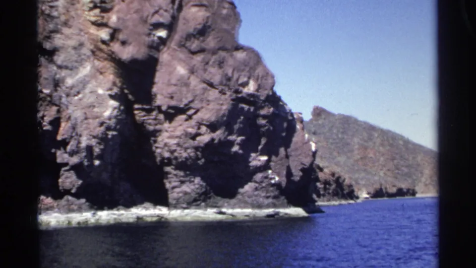 1975 8mm view near Guaymas showing a rocky cliff edge, white surf, blue sea, and a distant coastal hill.