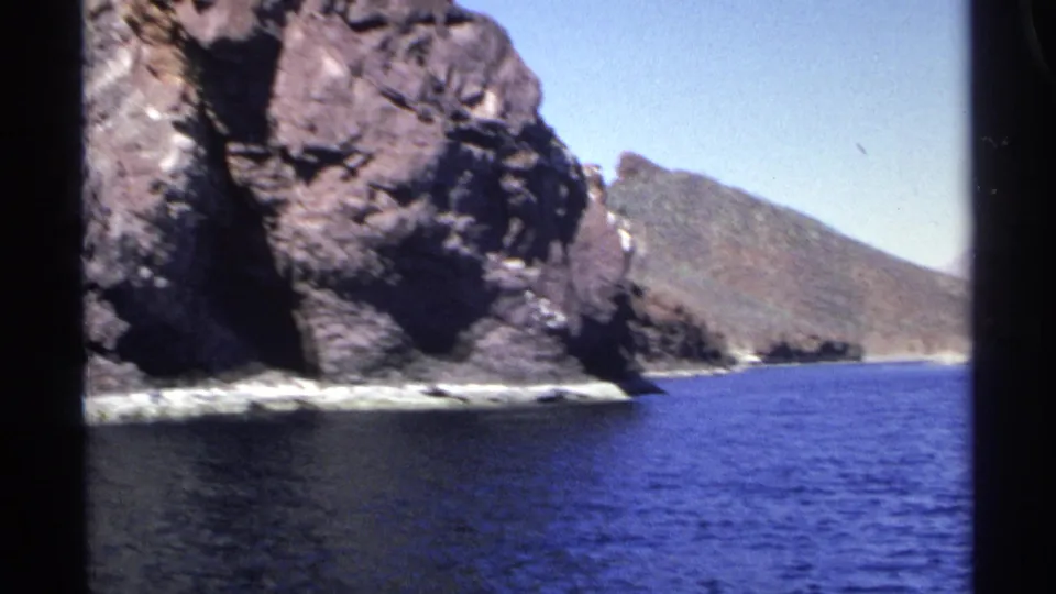1975 Guaymas coastal frame with dark rock formations, rippled blue water, white shoreline surf, and a distant hillside.