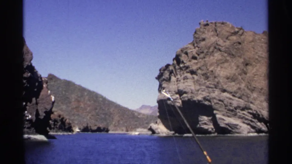 Boat-level 1975 Guaymas view of rocky coastline, blue water, and a fishing rod angled near the right edge.