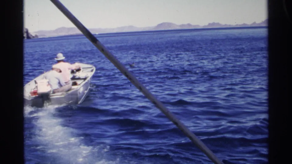 1975 Guaymas frame of a small motorboat carrying two men across blue water, leaving a wake under a distant mountain horizon.