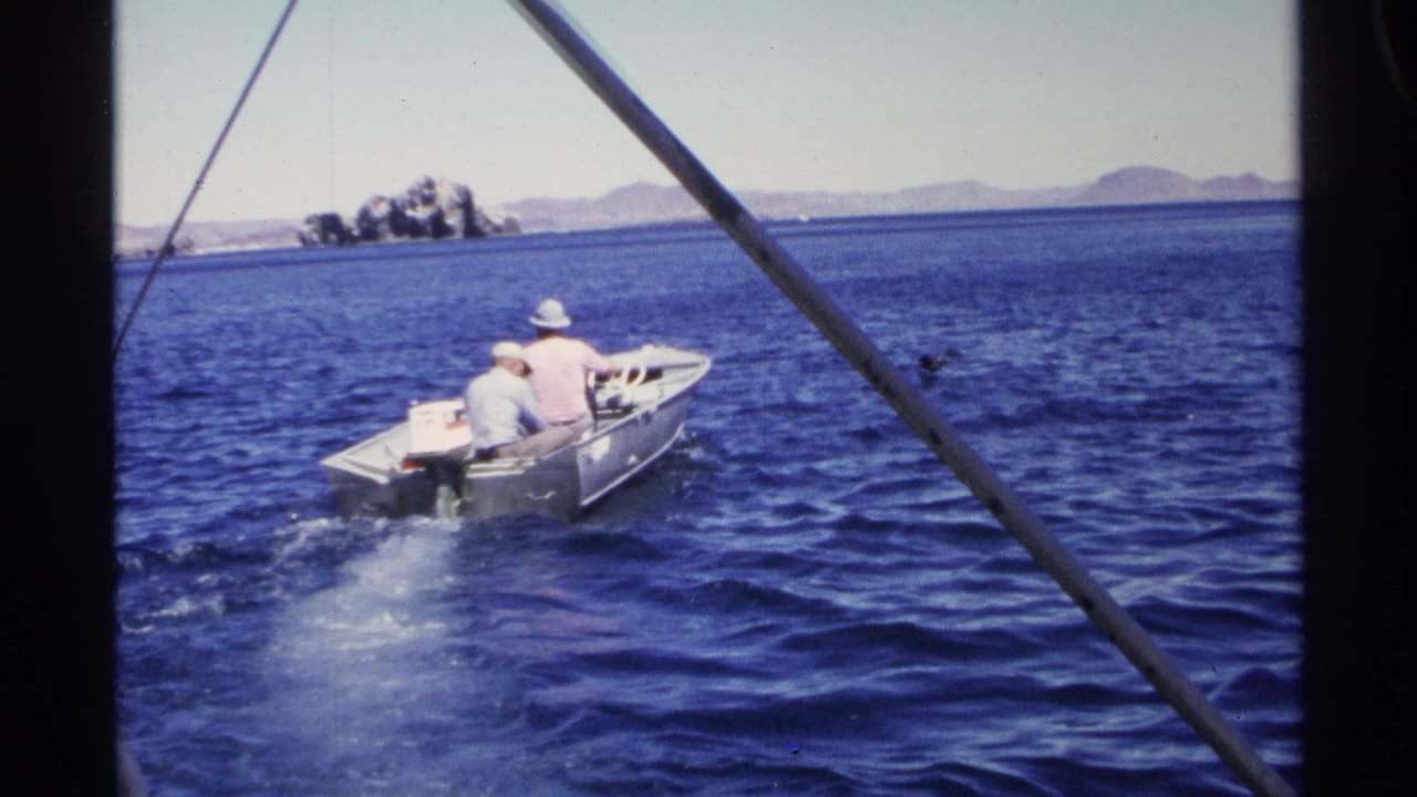 1975 Guaymas home-movie frame of a small motorboat carrying two people across deep blue water with a low mountainous coastline beyond.