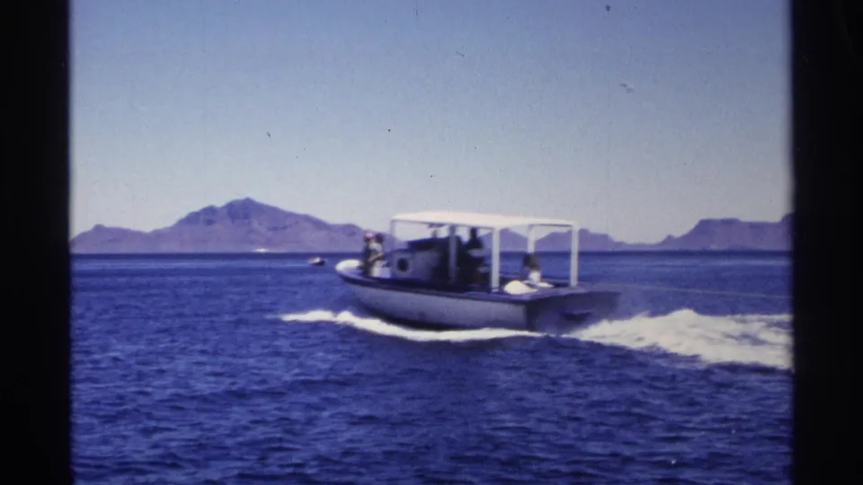 A covered motorboat with people aboard travels across Guaymas coastal water, leaving white wake below a clear sky and mountain horizon.