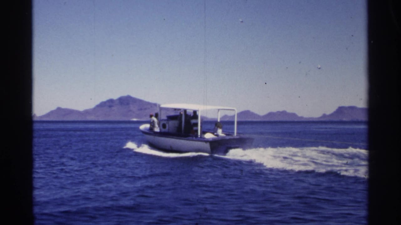 1975 Guaymas home-movie frame showing a small motorboat on blue water with distant mountains and bright daylight.