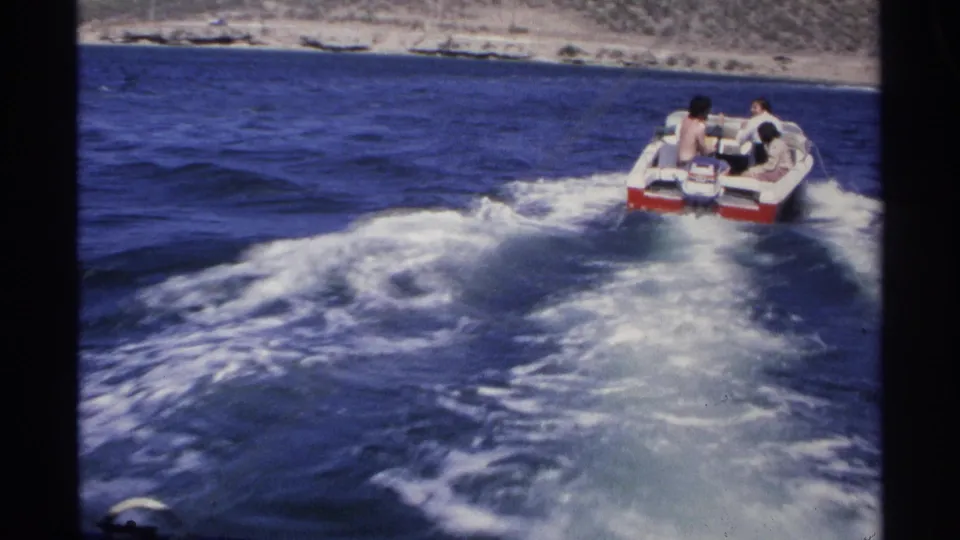 1975 Guaymas frame with passengers in a red-and-white speedboat moving away near a rocky coast and blue water.