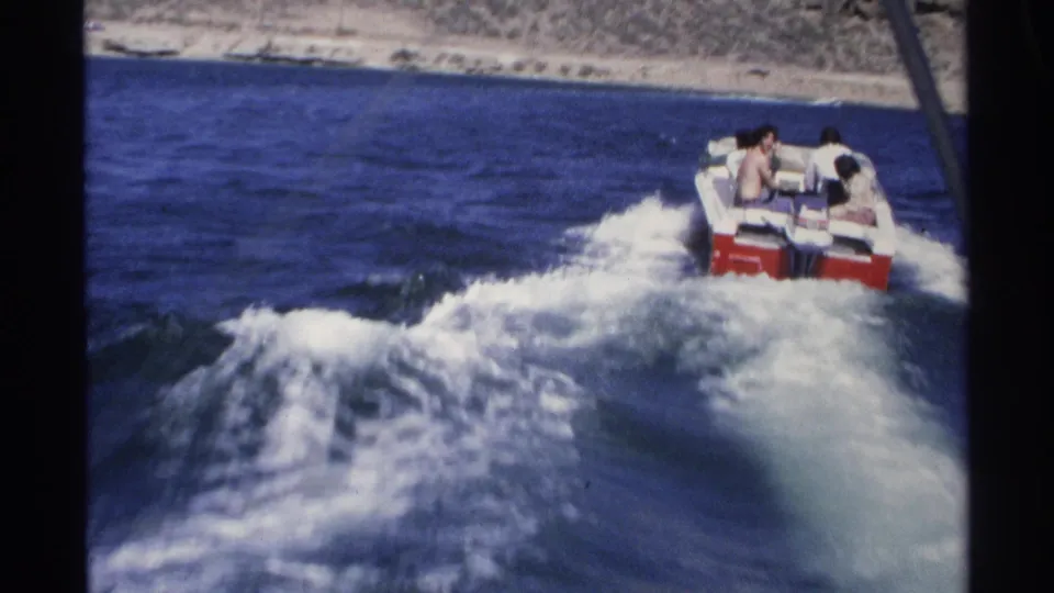 Red-and-white speedboat in 1975 Guaymas footage turning through choppy wake, with passengers visible against a rocky shoreline.