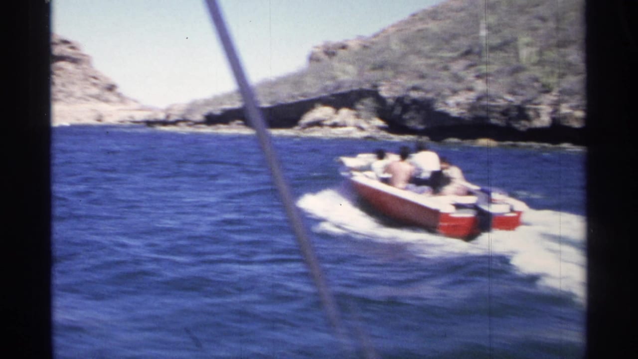 1975 Guaymas home-movie frame of a red motorboat with passengers crossing blue water beside a rocky, arid shore.