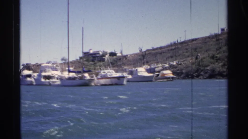 1975 Guaymas marina frame showing sailboats and motorboats moored near an arid hillside with small structures above the shore.