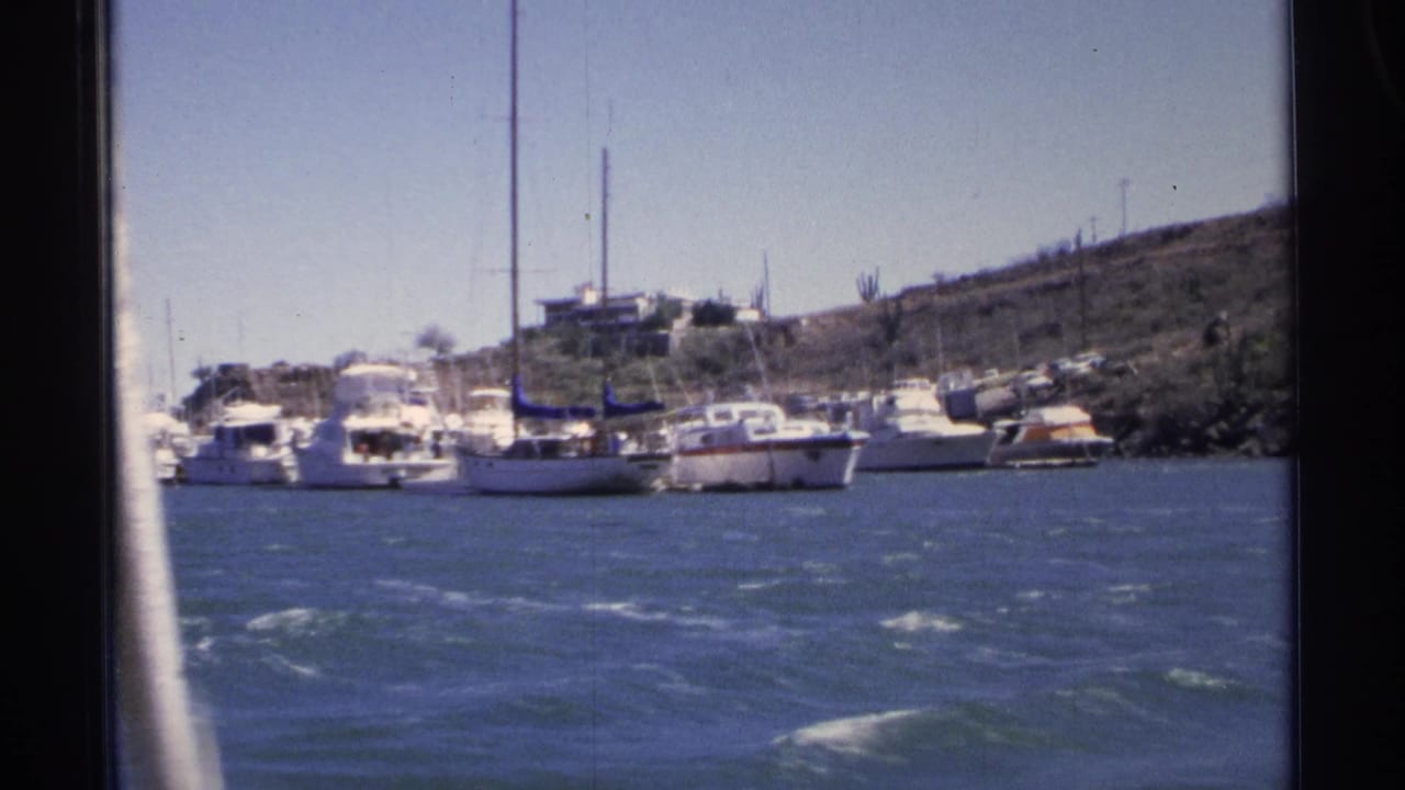 1975 Guaymas marina view from a moving boat, with moored yachts, sailboat masts, blue water, and a dry hillside in the background.