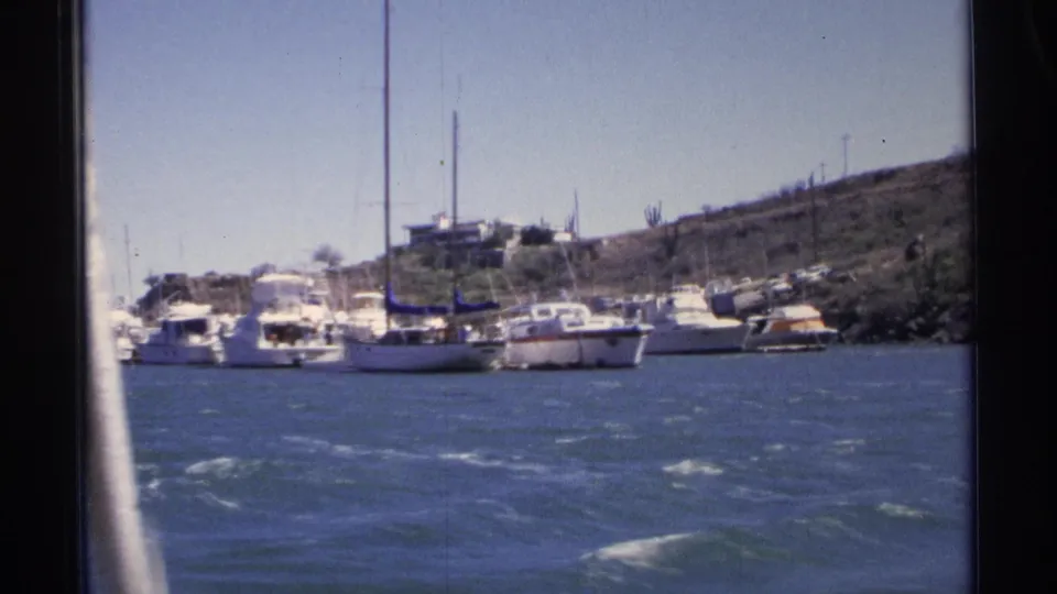 1975 Guaymas marina view from a moving boat, with moored yachts, sailboat masts, blue water, and a dry hillside in the background.