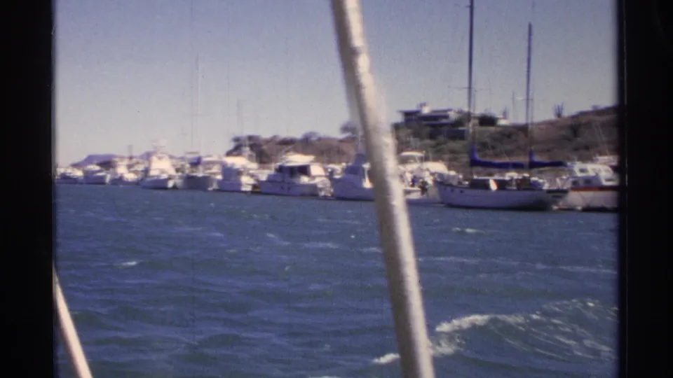 1975 home-movie frame of Guaymas harbor, showing clustered boats along the shore and a white mast from the filming boat.
