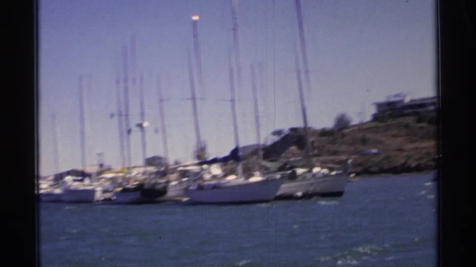 A tilted 1975 Guaymas marina frame with sailboat masts, moored vessels, blue water, and a low hillside behind the harbor.