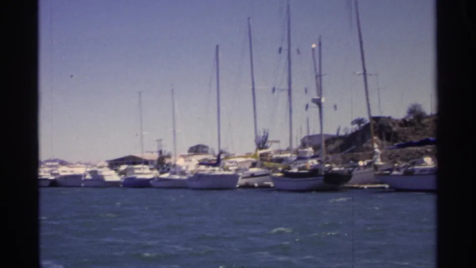 1975 8mm view of a Guaymas marina with rows of white boats, tall masts, shoreline structures, and blue water.