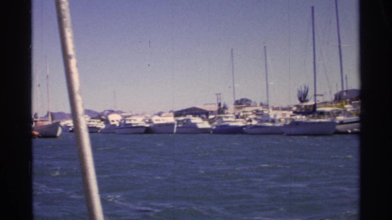 1975 8mm view from a boat in Guaymas, Mexico, looking across blue harbor water toward docked sailboats under a clear sky.