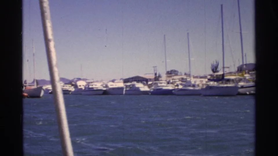 1975 8mm view from a boat in Guaymas, Mexico, looking across blue harbor water toward docked sailboats under a clear sky.