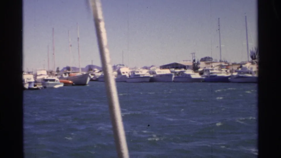 Guaymas marina scene from a moving boat, showing white sailboats, moored yachts, low shore structures, and 8mm film edges.