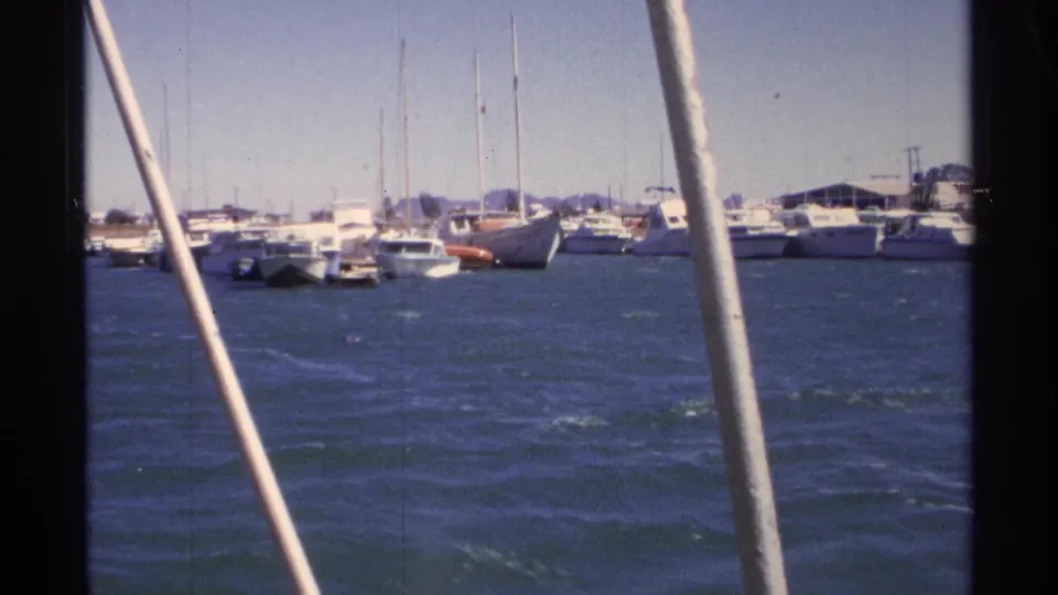 Moving 8mm boat perspective in Guaymas, with foreground rails framing docked boats, sailboat masts, and distant low land.
